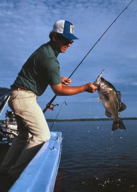 A Redondo bass is lifted from the lake for unhooking and releasing. While boats are acceptable for fishing, Russian-made Mockba motors were a disaster. Note the outboard behind the angler without its cowling, which was always removed for constant servicing required.